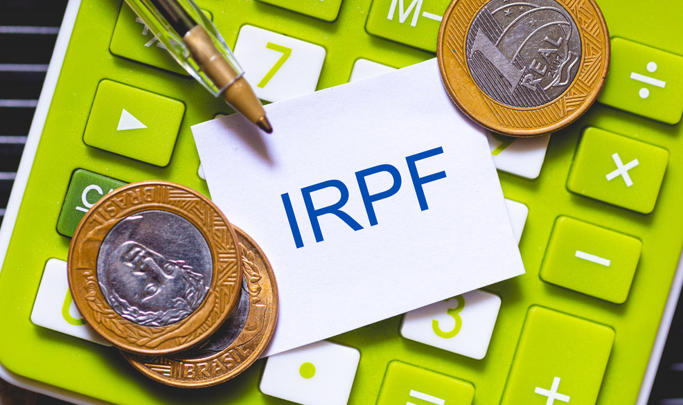 The initials IRPF written in blue color on a small piece of paper. Pen and Calculator in composition. Brazilian economy, income tax.
